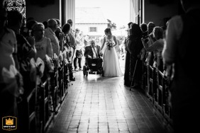 In a black and white shot inside a church in Meuse, France, the bride walks down the aisle with her grandfather in his wheelchair, the most important person in her life, capturing a moment of deep emotion and connection.